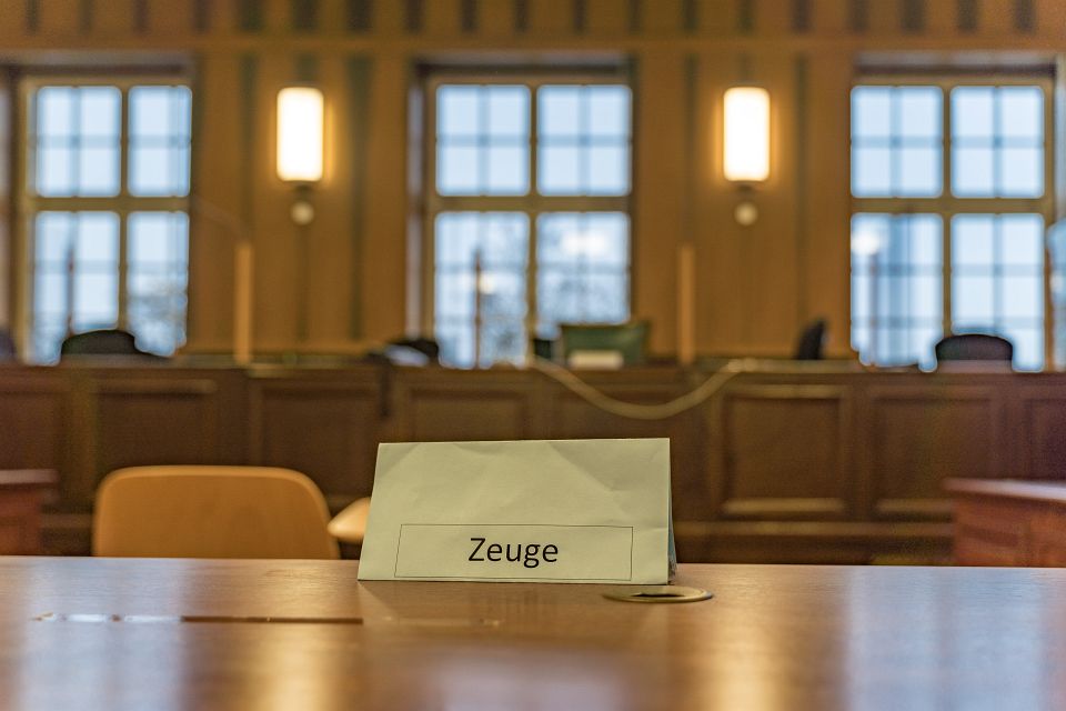 Ein Papierschild mit dem Aufdruck "Zeuge" steht auf einem Tisch in einem Gerichtssaal von 1870. Dahinter sieht man unscharf die leere Richterbank mit Fenstern dahinter.