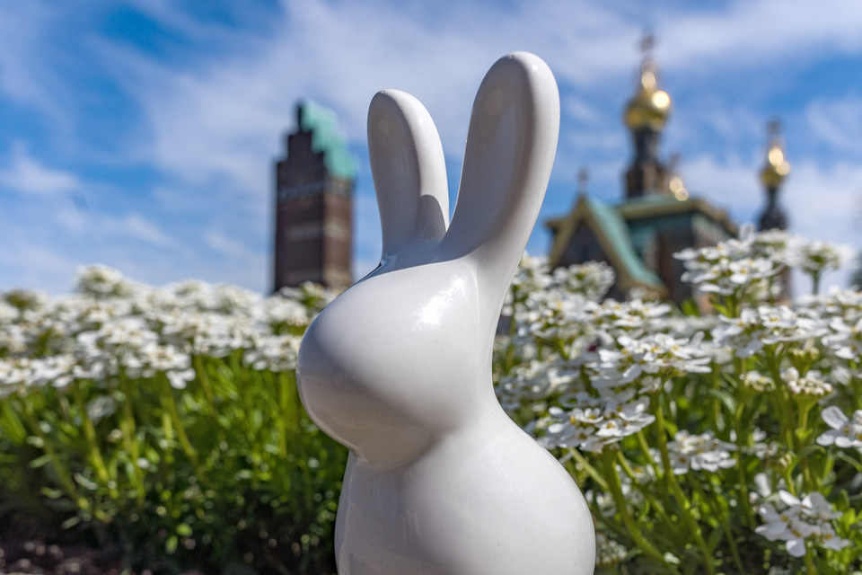 Ein nicht mal handgroßer weißer Hase aus Porzellan ohne Geicht steht vor kleinen blühenden Pflanzen. Im Hintergrund sieht man unscharf den Darmstädter Hochzeitsturm und die russische Kapelle auf der Mathildenhöhe.