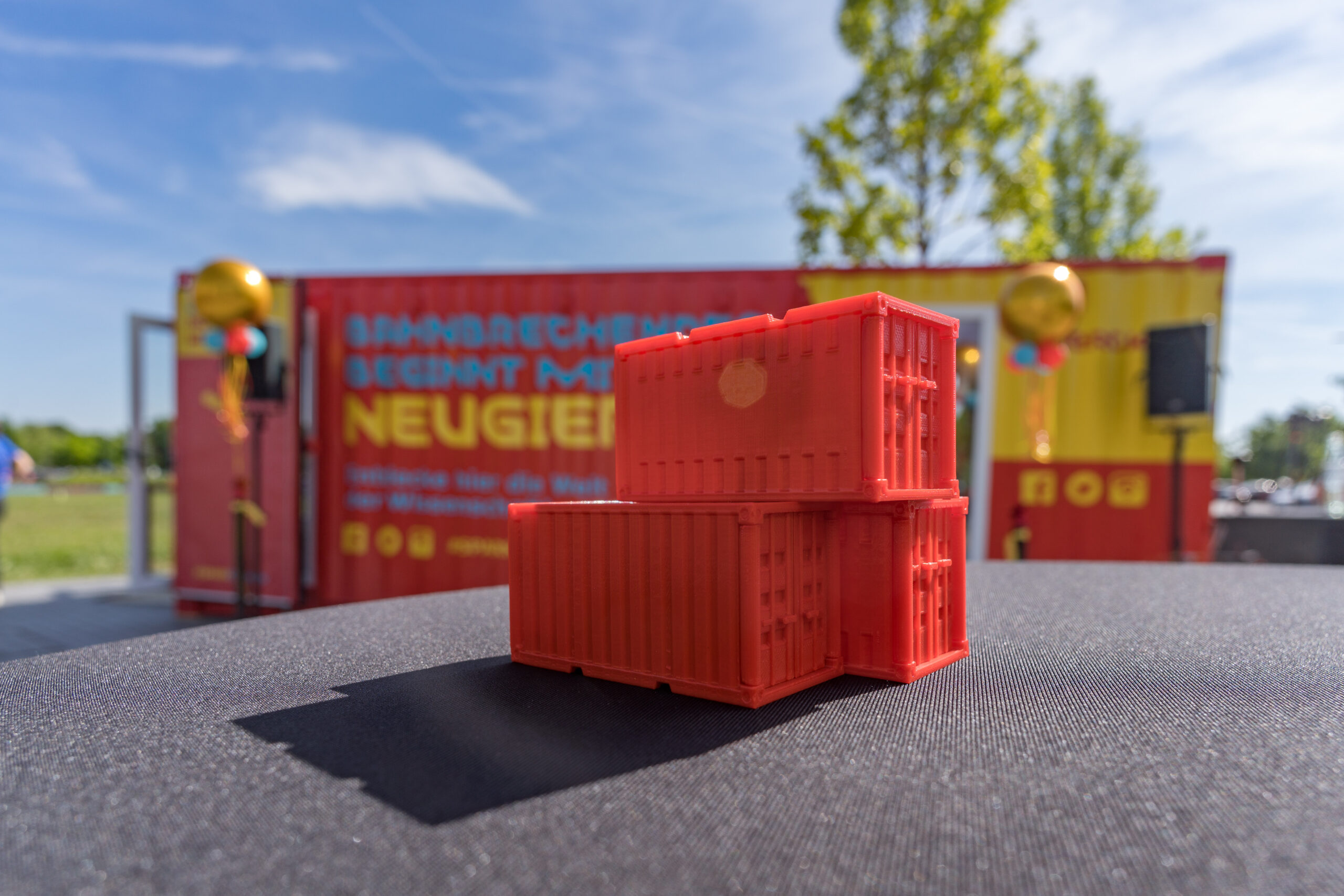 Drei kleine rote Spiezeug-Container stehen auf einem Tisch, dahinter steht ein echter Container, auf dem "Neugier" steht und in dem ein Merck-Schullabor drin ist.